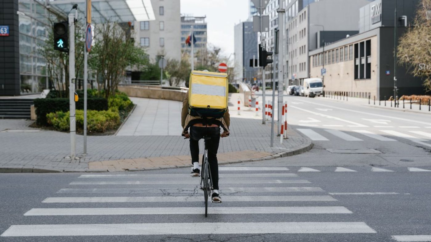 A courier crossing a city street by bicycle with an insulated delivery bag.